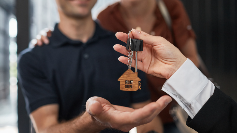 hand holding a house-shaped keychain with keys, being handed to a person, representing buying a home property