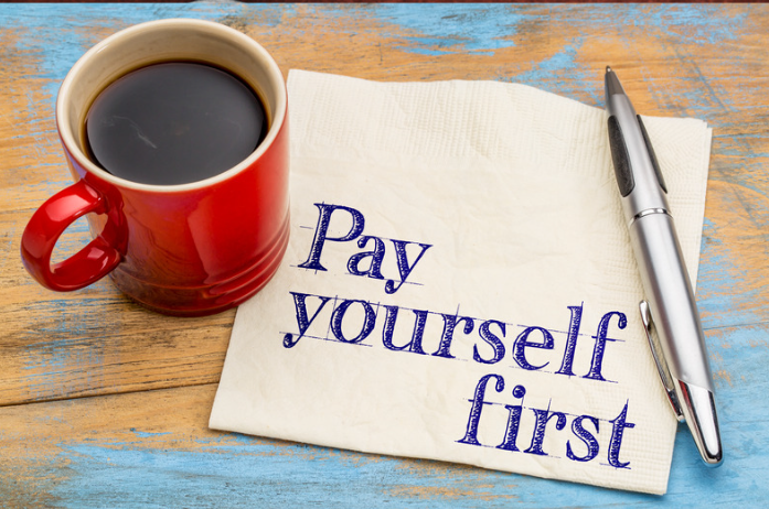 Red coffee cup and pen on a wooden table with a napkin that says 'Pay yourself first,'