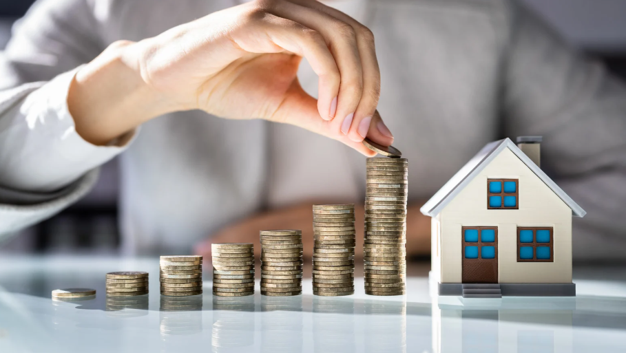 A hand stacking coins in ascending order next to a small house model