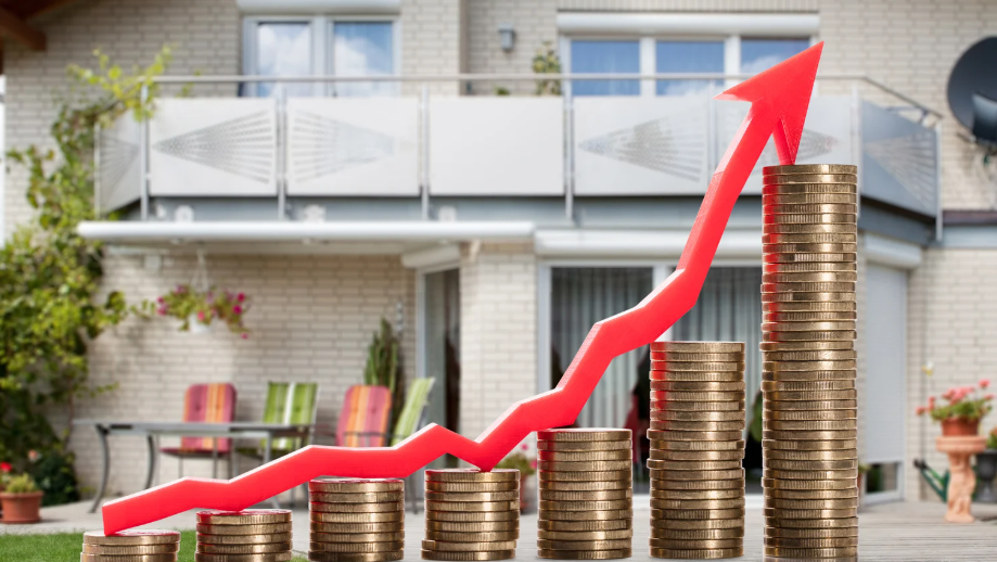 Stacks of coins with a rising red arrow in front of a modern house