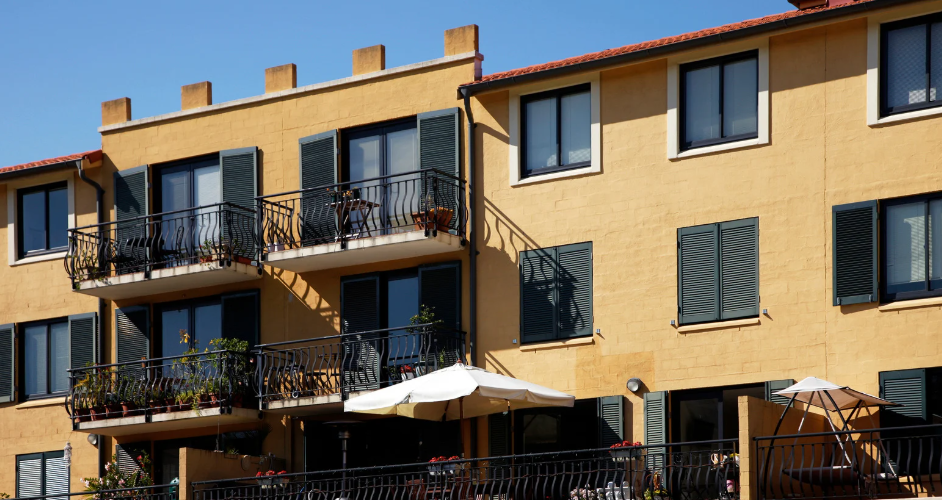 A yellow apartment building with black railings, balconies, and outdoor umbrellas set against a clear blue sky.