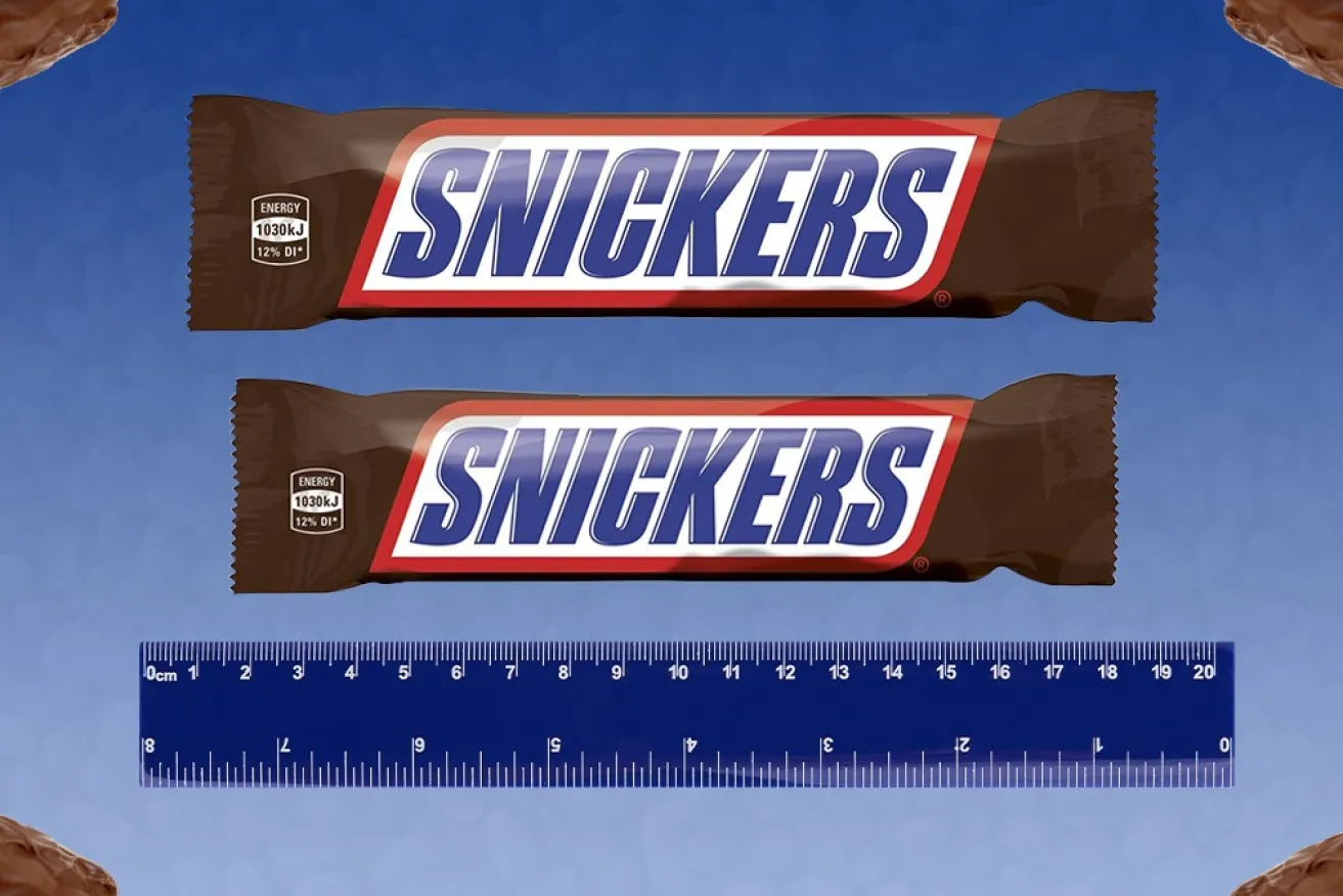 Two Snickers candy bars, one appearing larger than the other, placed above a ruler on a blue background with chocolate pieces in the corners.