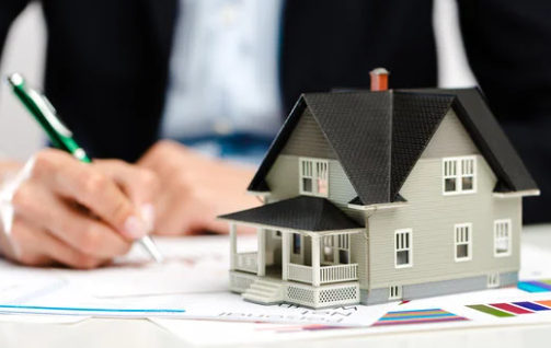 A person writes with a green pen beside a model house on a table covered with colorful charts and papers, focusing on real estate planning.