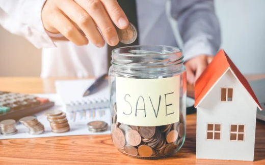 A person putting coins into a jar marked "SAVE" beside a small house, showing saving up to buy a home.