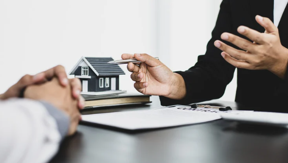 Two hands discussing real estate, with one pointing at a model house on a desk, showing a property consultation.