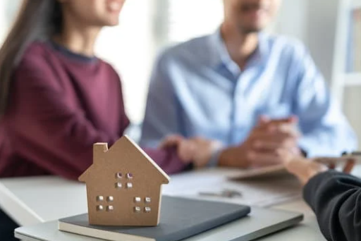 A cardboard house on a desk, with three people in the background, talking about real estate.