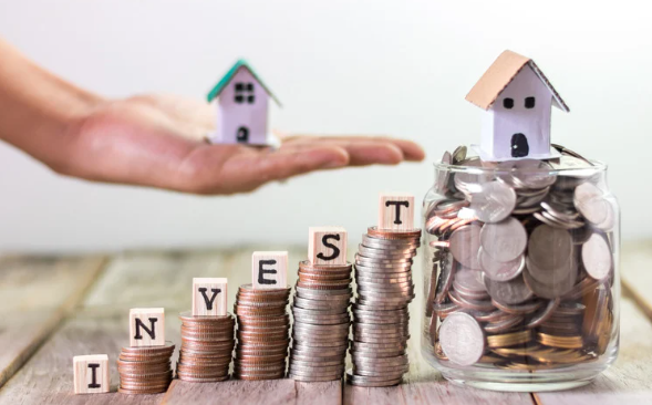 Stacks of coins spelling out the word "INVEST" beside a glass jar filled with coins and topped with a small house model, with another house model resting on a hand in the background.