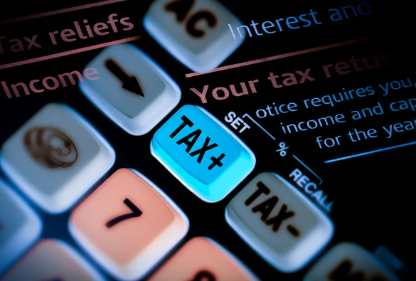 Close-up of a calculator with an illuminated "TAX+" button, surrounded by other keys like "SET", "TAX-", and numerical buttons, with a partially visible "Your tax return" notice in the background.