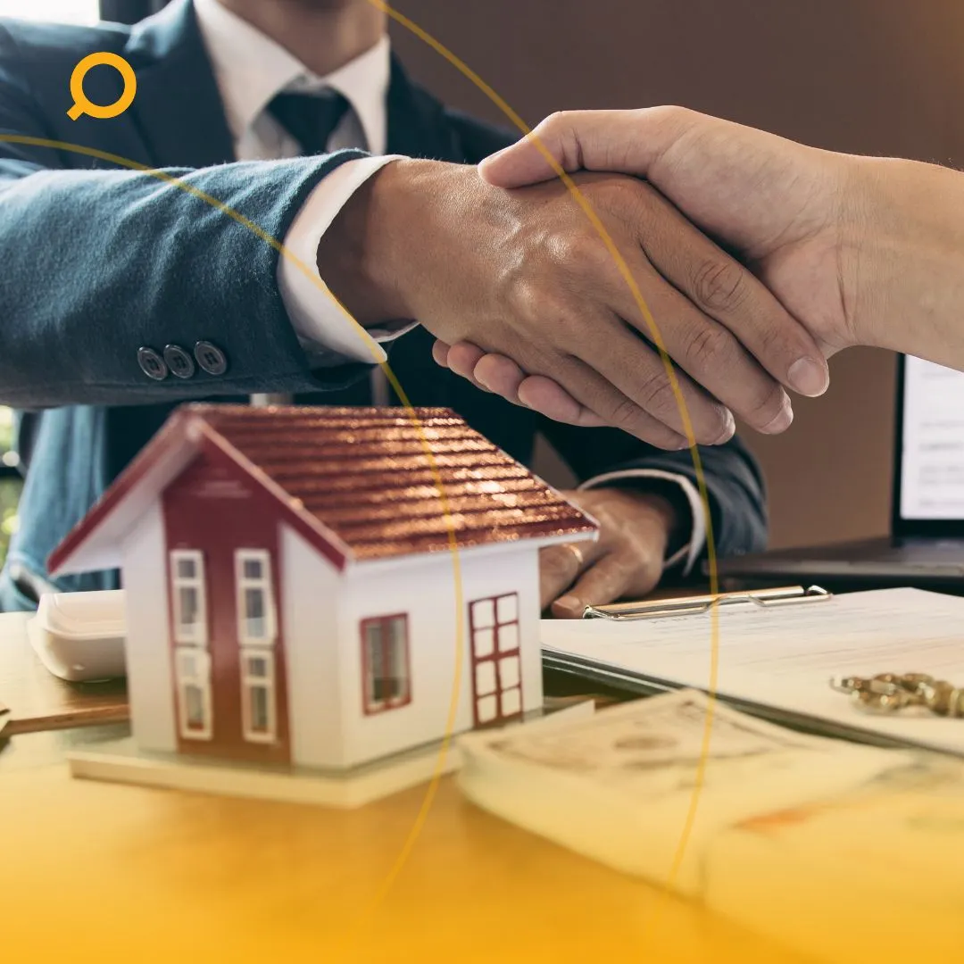 Two people shaking hands over a desk with a small house model and documents.