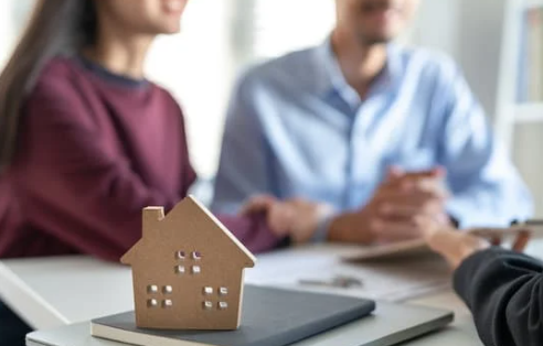 A wooden house model on a table in front of a couple shaking hands with a real estate agent.