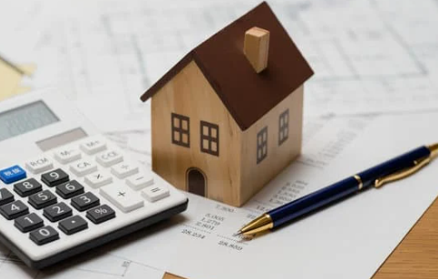 A wooden house model sits on financial documents, alongside a calculator and a blue pen.