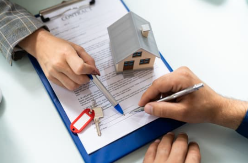 A close-up of two hands signing a contract with a model house and keys on the table.