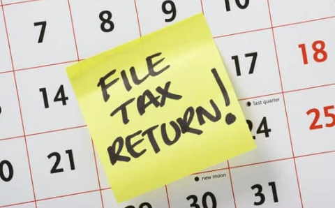 A close-up of a calendar with a bright yellow sticky note that reads "FILE TAX RETURN!" urging action on tax submission.