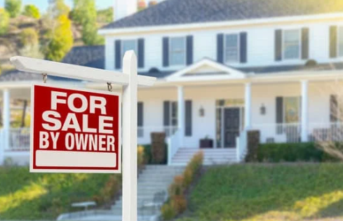 A "For Sale By Owner" sign stands in front of a charming white house with black shutters and a welcoming porch.