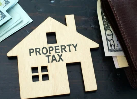 A wooden house shape labeled "PROPERTY TAX" beside cash and a wallet on a dark surface.