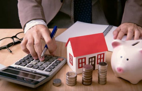 A person calculates finances with a calculator, surrounded by a model house, piggy bank, and stacks of coins, suggesting budgeting for housing.