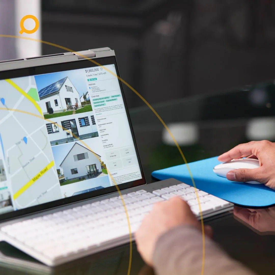 A person using a computer, viewing property listings with images and a map on the screen, while navigating with a mouse.