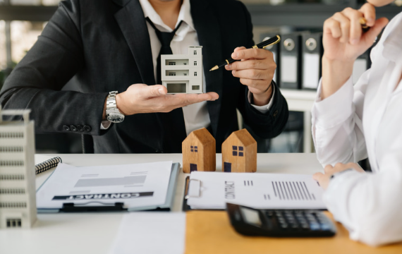 A businessman presents a model house while discussing real estate with a colleague, with documents and miniature houses on the table.