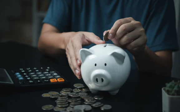 A person adding a coin to a white piggy bank, surrounded by scattered coins and a calculator on a dark table.