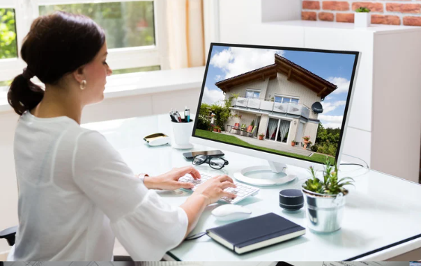 A woman works at a desk, viewing a large monitor displaying a photo of a modern house surrounded by greenery.