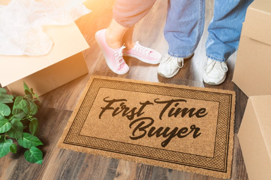  Doormat with "First Time Buyer" text, surrounded by moving boxes and two people standing side by side.