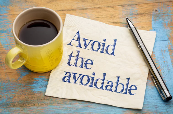 Yellow coffee mug, silver pen, and napkin with the phrase 'Avoid the avoidable' written on it, placed on a rustic wooden table.