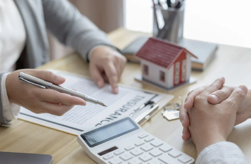 Close-up of two people reviewing insurance documents with a miniature house, calculator, and keys on the table—representing property insurance planning and financial discussions.
