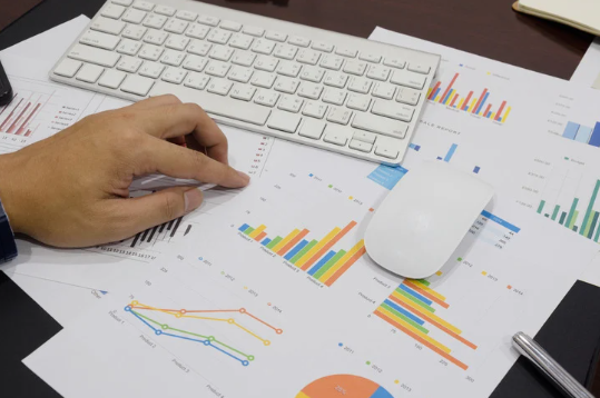 Person analysing colourful financial graphs on printed sheets with a white computer keyboard and mouse, representing investment strategy and data analysis.