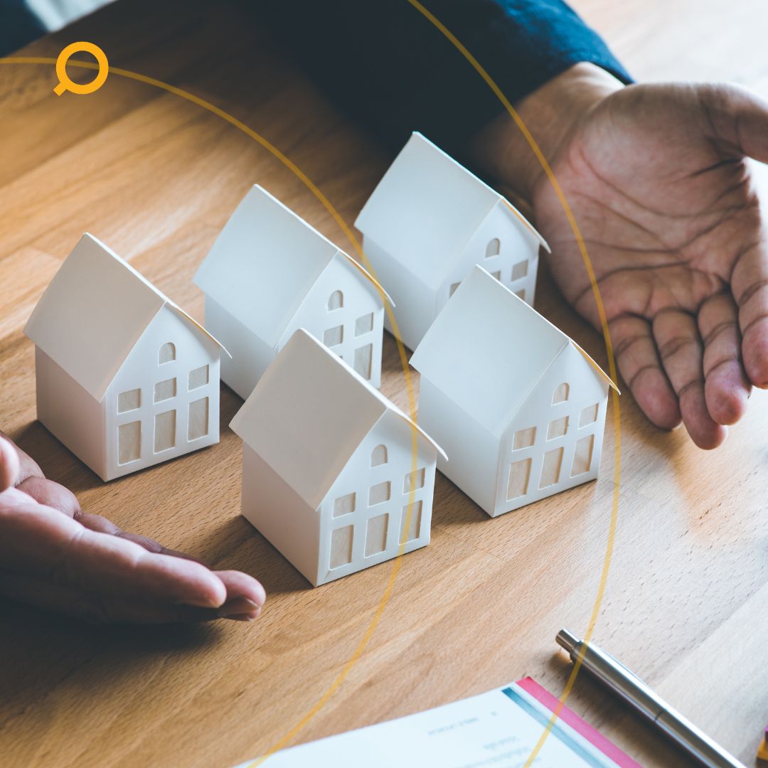 Hands surrounding miniature house models on a wooden desk, representing property investment or real estate portfolio planning.