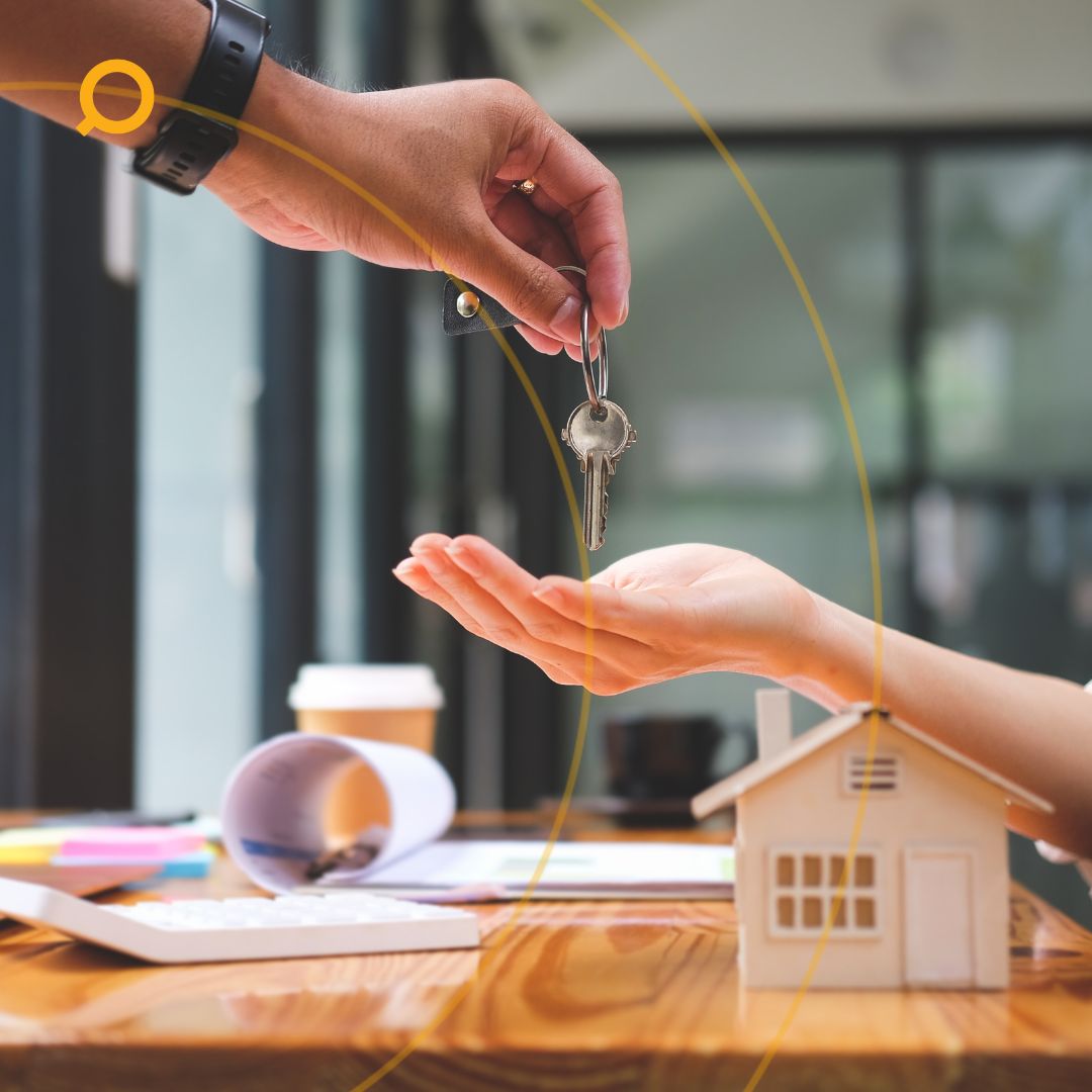 A person handing over house keys to a new homeowner across a desk with property documents, coffee, and a miniature house model.
