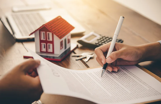 Person signing real estate contract with a model house, calculator, and keys in the background, representing a property purchase or investment agreement