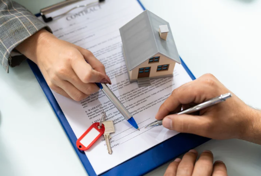 Homebuyer signing homeownership contract with house model and property keys on table.