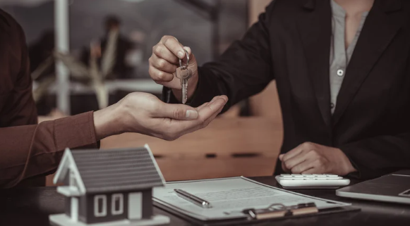 Real estate agent handing over house keys to a new homeowner, with a contract, pen, and model house on the desk symbolising property purchase or settlement.