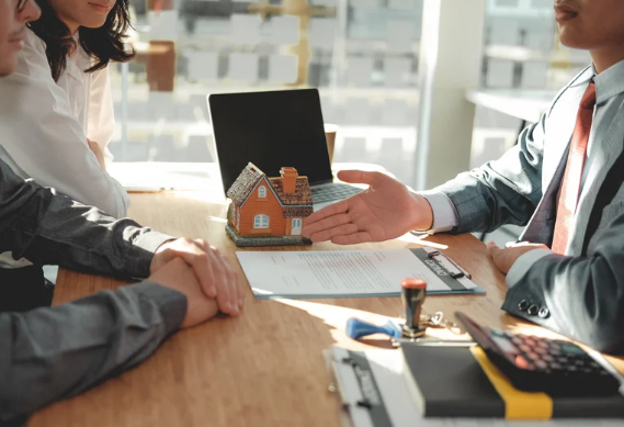 Real estate agent presenting a miniature house to clients during a property purchase meeting, with contract and calculator on the table.