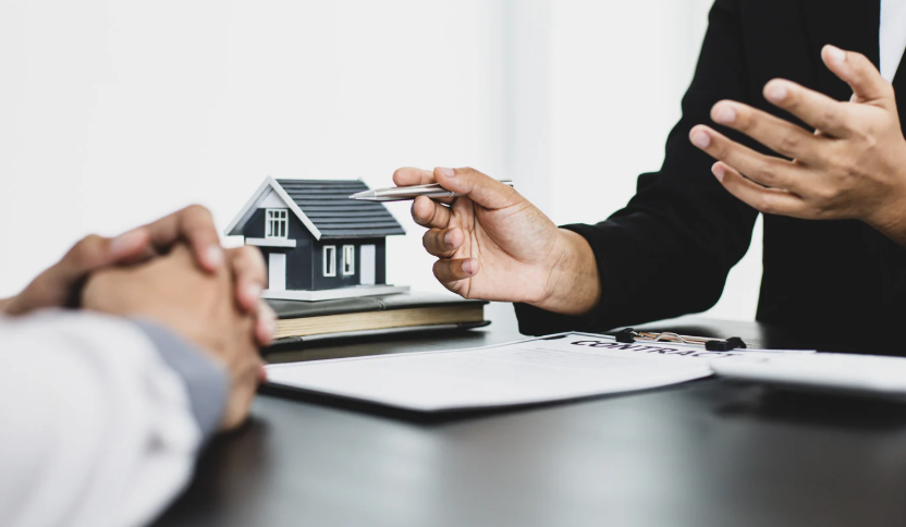 Real estate agent discussing property investment with client, pointing at house model and contract paperwork.
