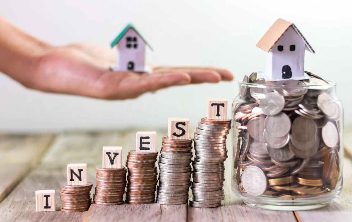 Jar filled with coins topped by a miniature house, stacked coins spelling INVEST, and a hand holding a small house, representing property investment growth.