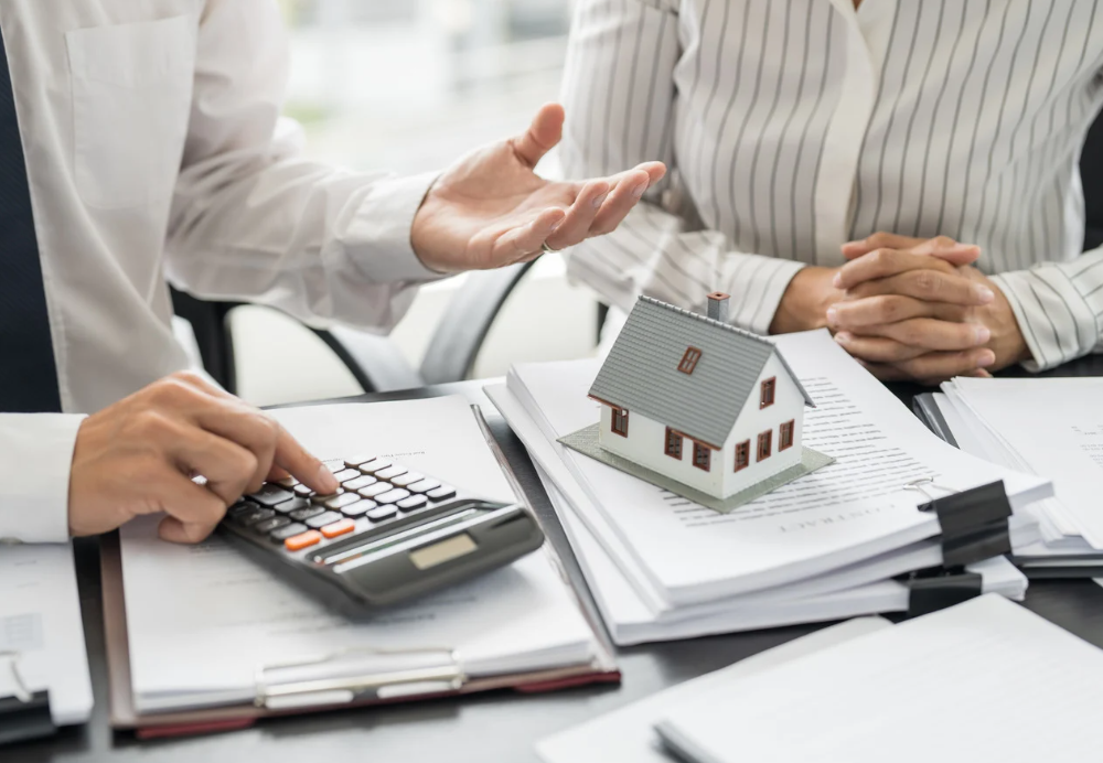Two people discussing property investment with a miniature house and calculator on the table, representing home loans and real estate strategy.