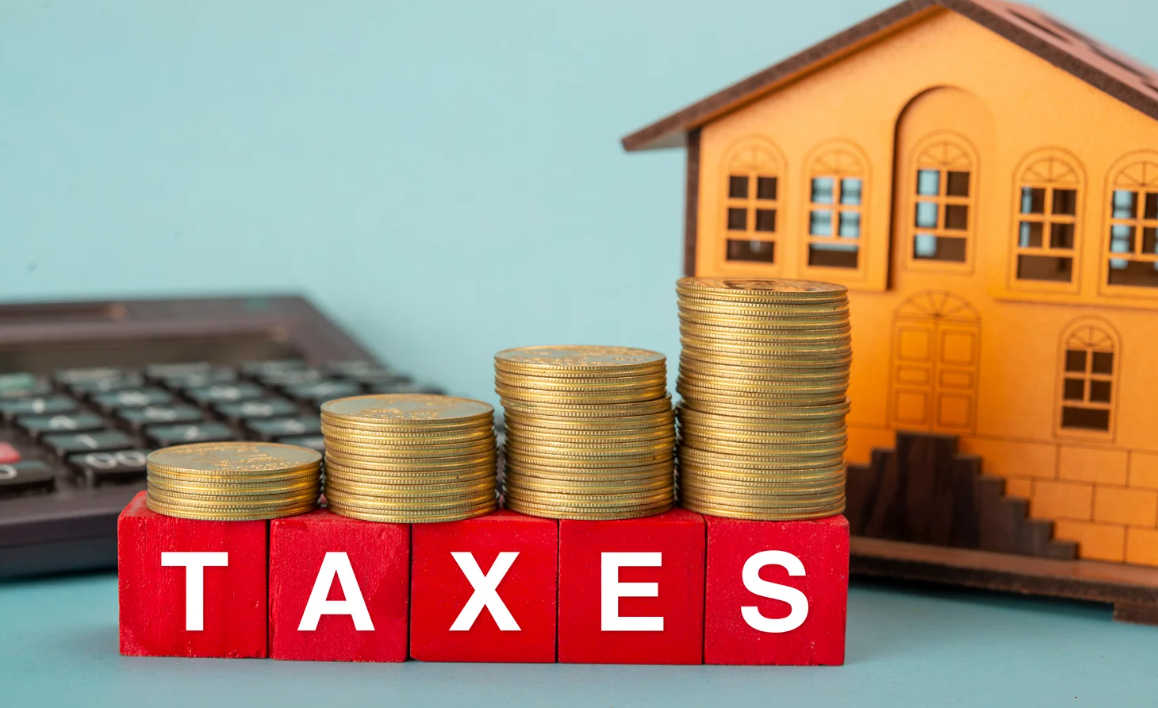Stacked gold coins on red blocks spelling “TAXES,” with a small wooden house and calculator in the background, representing property tax and investment finance.
