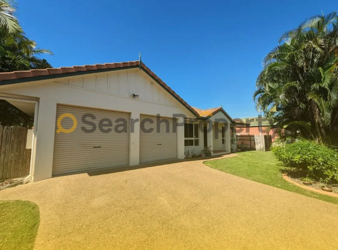 Front view of a single-storey suburban house with a double garage, gabled roof, and well-maintained driveway and lawn, surrounded by tropical greenery under a clear blue sky.